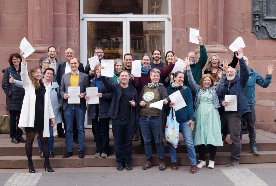 Eine Gruppe an Menschen steht vor einem Rathaus in Deutschland und halten feiernd ein Blatt Papier in die Kamera