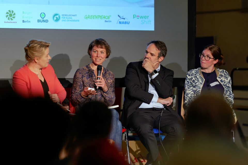 Vier Personen sitzen auf einer Bühne und nehmen an einer Podiumsdiskussion teil. Sie sind in ein Gespräch vertieft und halten Mikrofone in den Händen. Die zweite Person von links spricht gerade und gestikuliert mit der rechten Hand, während die anderen aufmerksam zuhören.